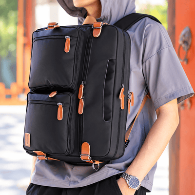 A person wearing a gray short-sleeve shirt carries a black multi-compartment backpack with tan leather-like zipper pulls and straps in an outdoor setting with warm-toned architectural elements.