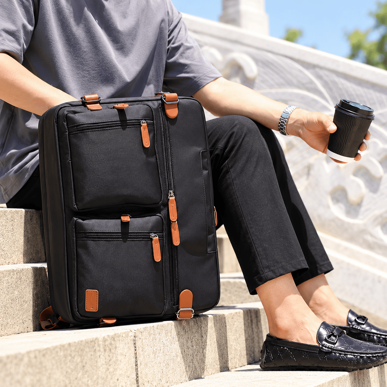 A person sitting on outdoor stone steps beside a black rolling suitcase with multiple zippered compartments and tan leather-like zipper pulls, while holding a takeaway coffee cup.