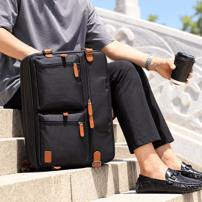 A person sitting on outdoor stone steps beside a black rolling suitcase with multiple zippered compartments and tan leather-like zipper pulls, while holding a takeaway coffee cup.