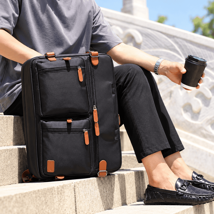 A person sitting on outdoor stone steps beside a black rolling suitcase with multiple zippered compartments and tan leather-like zipper pulls, while holding a takeaway coffee cup.