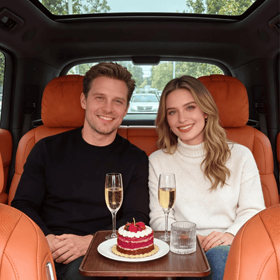 A man and a woman sitting in the back of a luxury vehicle with orange leather seats, with a small red velvet cake on a tray table between them, flanked by two champagne glasses.