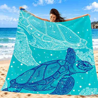 A woman on a sandy beach holds up a large turquoise towel or blanket featuring two white and blue sea turtle designs with intricate mandala-style line art.