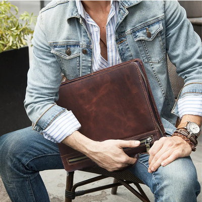 A man in a denim jacket and jeans holds a brown leather briefcase or document pouch with a zipper closure while sitting on a chair.