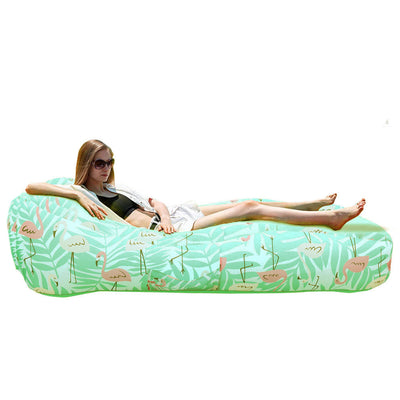 A woman reclines on a mint green inflatable lounger with a tropical flamingo and palm leaf print. A small inset at the top right shows the same item carried on a backpack.