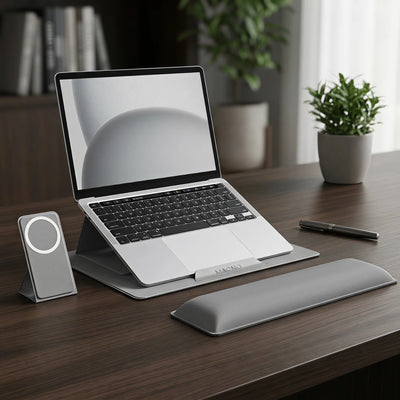 A silver laptop stand and matching wrist rest arranged on a dark wood desk, with a laptop placed on top. A small gray device with a circular light is visible to the left, and a pen lies to the right.