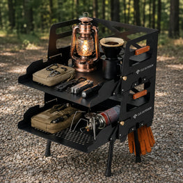 A black outdoor camping cooking cart with two shelves and side handles, shown on a gravel path in a forest. The cart is styled with camping gear including a lit copper lantern, a coffee maker, a folding knife, a small pouch, and a stove fuel canister.