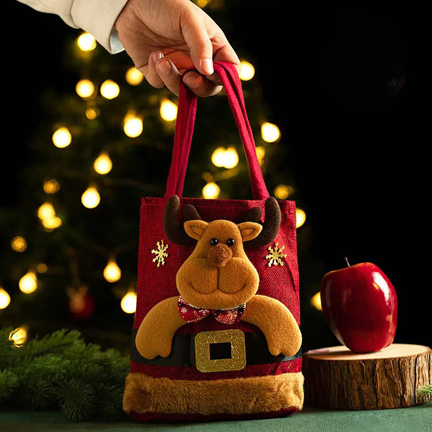 A red felt Christmas tote bag with a plush reindeer face and Santa-style belt, held by a hand in front of a blurred Christmas tree with warm lights. A red apple sits on a wood slice beside it.