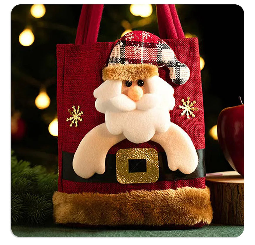 A red fabric tote bag decorated with a plush Santa Claus face, a plaid Santa hat, gold snowflake details, and a faux fur trim at the bottom, shown against a festive blurred holiday background.