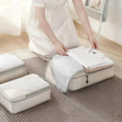 A woman in a white dress packs a white shirt into a light gray mesh zippered travel organizer bag on a textured rug. Two more closed travel bags are visible nearby in a softly lit indoor setting.
