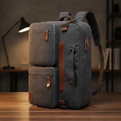 A dark gray canvas laptop backpack with brown leather accents, standing on a wooden table in a dimly lit room with a lamp and shelves in the background.