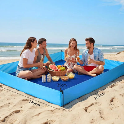 A blue inflatable beach blanket with a built-in umbrella frame is set up on a sandy beach with four people sitting inside, holding drinks and enjoying a picnic. The image includes dimension labels showing the blanket's size (145cm by 185cm by 25cm).