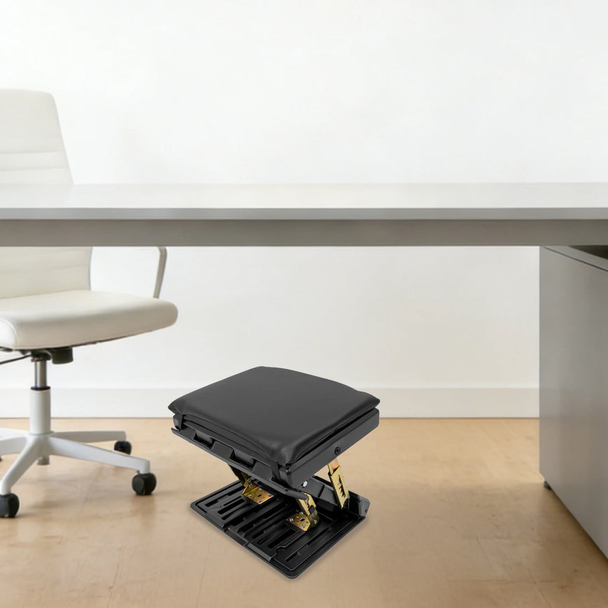 A black office footrest with gold-toned metal supports, positioned under a light gray desk in a minimalist office setting with a white chair and light wood floor.