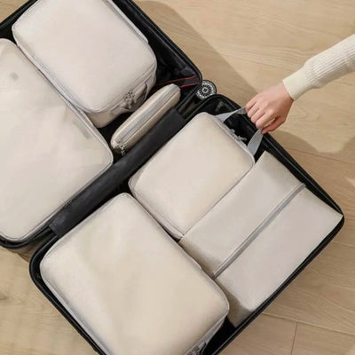A set of off-white packing cubes and a small zip pouch organized inside an open black suitcase on a light wood floor, with a person's hand and sleeve visible adjusting the contents.