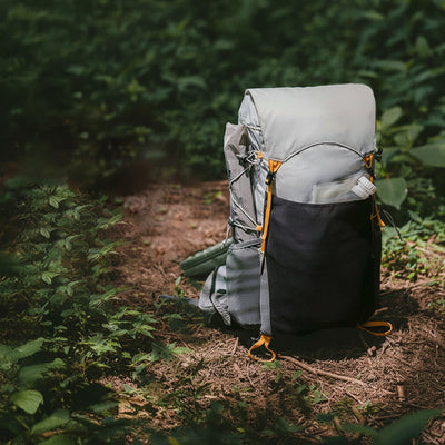 A Naturehike hiking backpack in light gray and black with bright orange accents, shown on a forest path with foliage and sunlight filtering through.