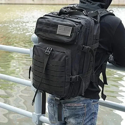 Person wearing a black tactical backpack with an American flag patch, standing by a body of water.
