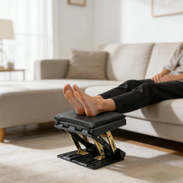 A black footrest with gold-toned metal supports and a padded top, shown with a person's bare feet resting on it in a living room setting.