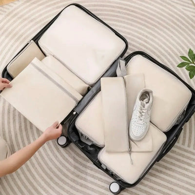 A black hard-shell suitcase opened on a striped rug, filled with multiple cream-colored packing cubes and a white sneaker. A person's hands are visible on the left, adjusting one of the packing cubes.