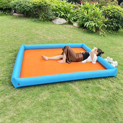 A woman lies on an inflatable pool or slide mat outdoors on a grassy lawn. The mat has a bright orange center and a blue inflatable border, with shrubs and trees in the background.