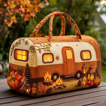 A decorative camping-themed handbag shaped like a vintage camper trailer, placed on a wooden table outdoors with autumn foliage in the background. The bag features warm orange and cream panels, floral-patterned handles, and printed autumn motifs including pumpkins, lanterns, campfires, and trees.