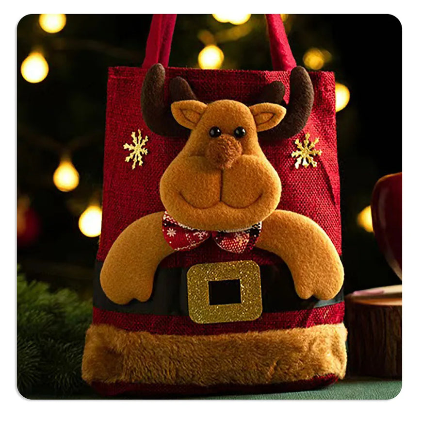 A festive Christmas-themed tote bag featuring a plush reindeer face and antlers, styled like Santa's belt with a gold buckle, red fabric body, and gold snowflake accents, set against a blurred holiday background with warm lights.