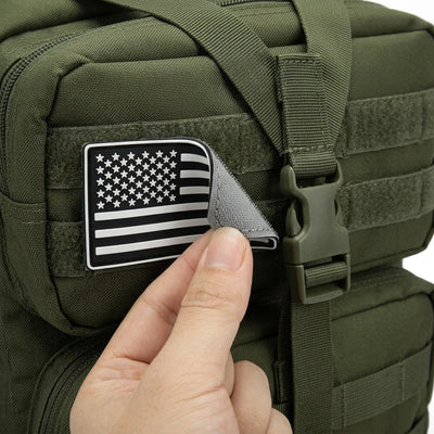 A close-up of an olive green tactical backpack with a black and white American flag patch on the front, being peeled off by a hand.