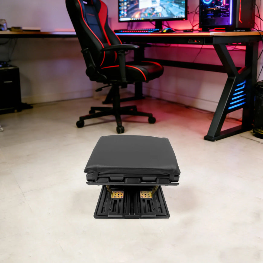 A black gaming footrest with a padded top and dual metal pedals, positioned on a light-colored floor in front of a desk setup with a black and red gaming chair.