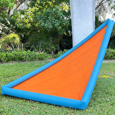 An inflatable pool slide with a bright orange inner surface and blue inflatable walls, set outdoors on a green lawn with trees and a concrete post in the background.