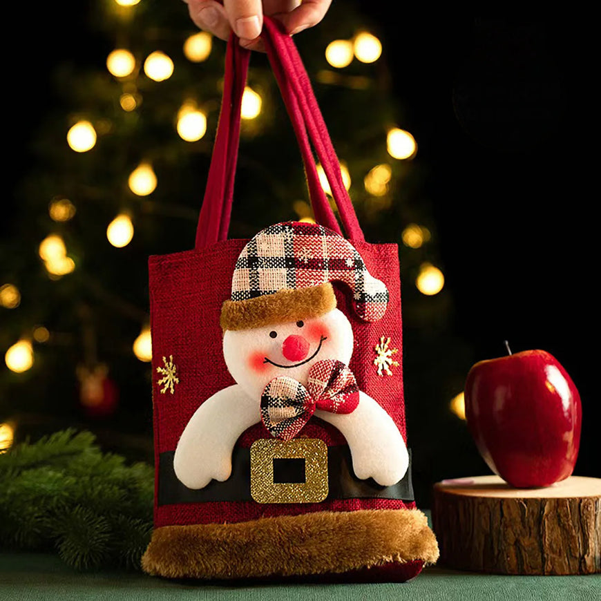 Decorative gift bag with a snowman design held by a hand, Christmas tree and apple in the background.