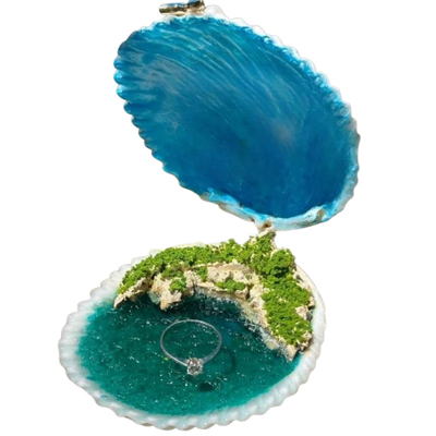 A seashell-shaped ring box shown open, with a miniature tropical island scene inside. The shell interior is bright blue on the outside and turquoise on the inside, with a green mossy island, white rocks, and a clear ring displayed on the bottom.