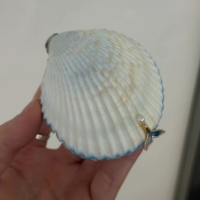 A hand holding a white scallop shell pendant with a small blue and gold finial charm and a tiny pearl accent, shown against a plain light background.