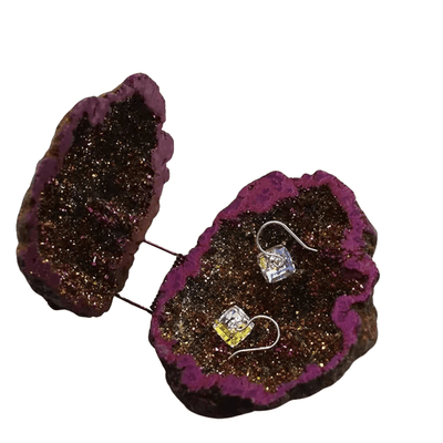 A pair of small silver hook earrings with clear and yellow square gemstones, displayed on a split geode-style stand made of dark glittery rock with a purple interior, against a plain white background.