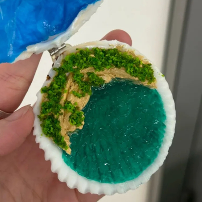 A hand holding a miniature shell-shaped object that resembles a beach or landscape scene, with bright green grass-like texture, tan ground, and a teal ocean-like area. A blue textured piece is attached at the top.
