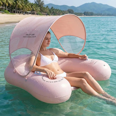 A woman wearing a white bikini sits in a pink inflatable pool float with a curved sunshade canopy, floating in clear turquoise ocean water with a sandy beach and mountains in the background.