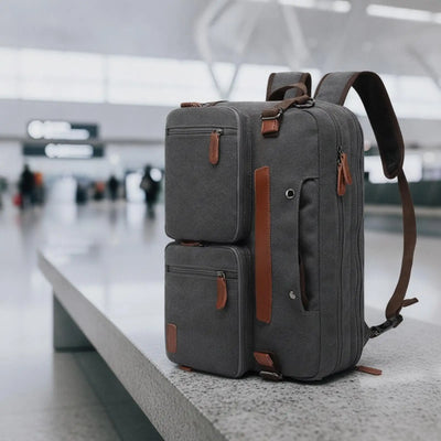 A dark gray canvas backpack with brown leather accents sits on a concrete ledge in a blurred airport terminal.