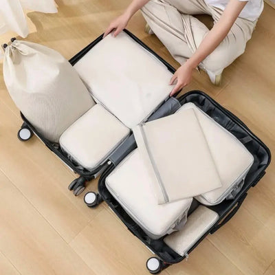 A person packing a black suitcase with multiple off-white fabric packing cubes and a matching drawstring bag on a light wood floor.