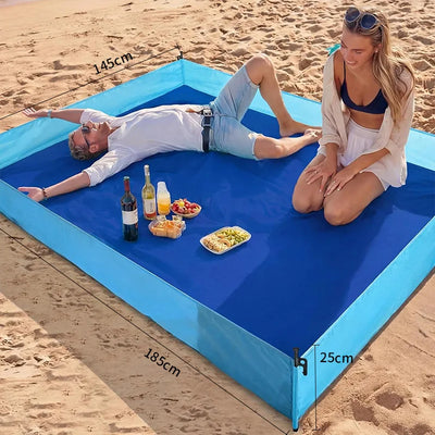 Two people on a blue inflatable mat with food and drinks on a sandy beach.