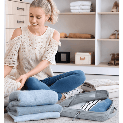 A woman sits on the floor in a closet area, folding light blue garments beside an open gray suitcase with folded clothes inside.