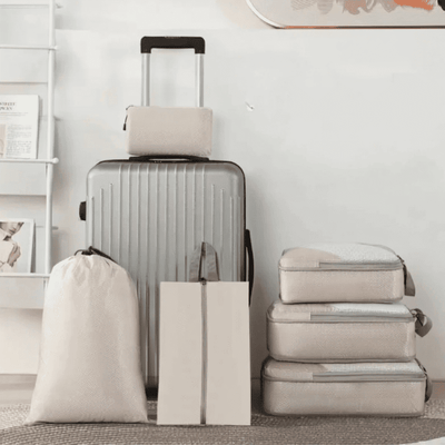 A travel luggage set arranged against a light wall, including a silver hardshell suitcase with the telescoping handle extended, three beige fabric storage bags, and two zippered packing cubes stacked on the right.
